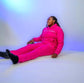 Taller Than LIfe Collection model showcasing ENDLESS HEIGHTS tracksuit set in vibrant pink, sitting on a white surface.